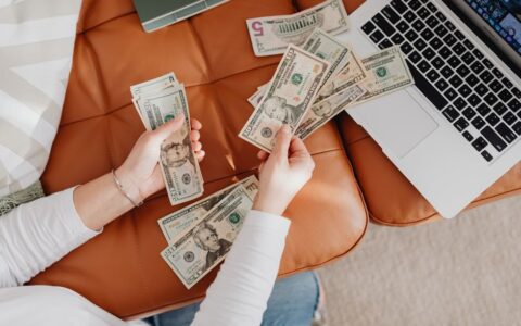 A person counts US dollar bills on a couch with a laptop, symbolizing financial planning.