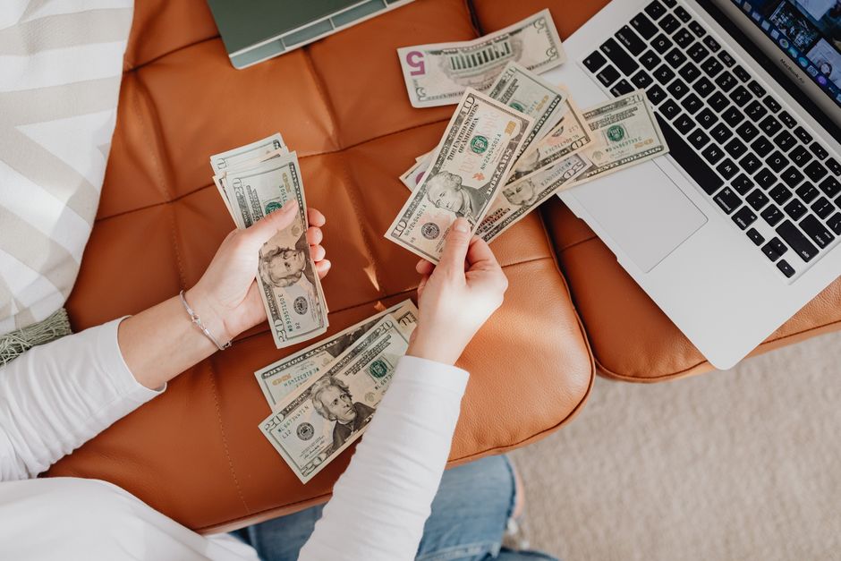 A person counts US dollar bills on a couch with a laptop, symbolizing financial planning.