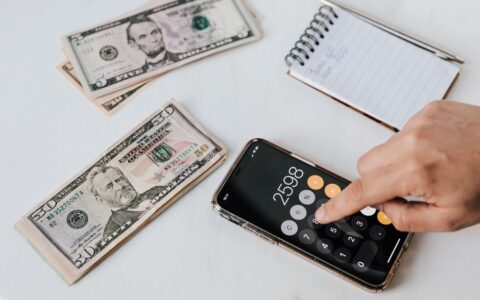 From above of crop anonymous economist calculating total amount of income using calculator app on cellphone near piles of greenbacks and notebook with pen