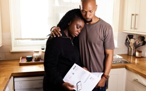 A couple embraces in their kitchen while going through important documents, reflecting concern and support.