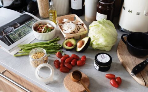 From above of various organic vegetables chopped ingredients near cutting board kitchenware appliances and trendy magazine placed on gray countertop in kitchen at home