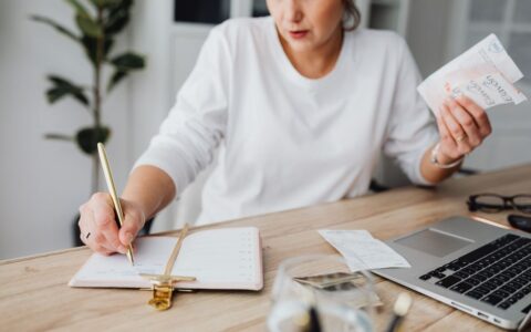 Woman managing finances, writing on notepad, and holding receipts at a desk.