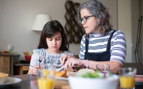 A mother helps her daughter cut food during breakfast at home, fostering family bonding indoors.