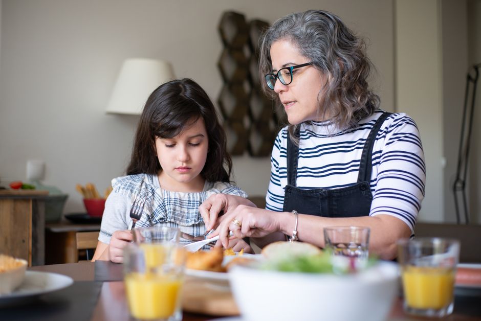 A mother helps her daughter cut food during breakfast at home, fostering family bonding indoors.