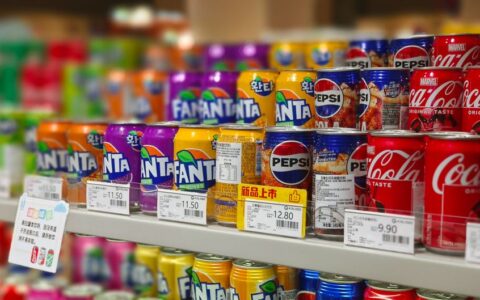 Colorful soda cans including Fanta, Pepsi, and Coca-Cola arranged on supermarket shelves, price tags visible.