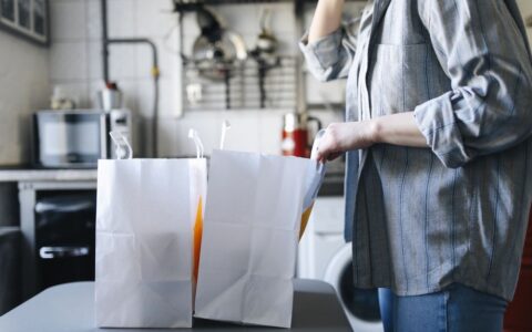 Woman in casual attire handling paper bags for delivery service in a kitchen setting.