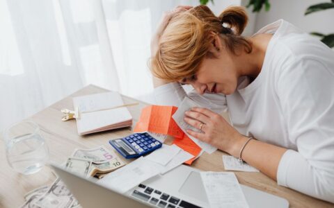 A woman overwhelmed with finances, surrounded by bills, calculator, and cash at home.