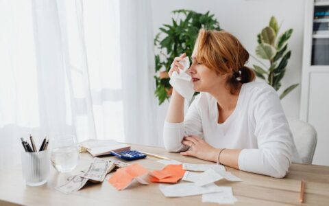 A woman is overwhelmed by bills and finances, sitting at a desk indoors.