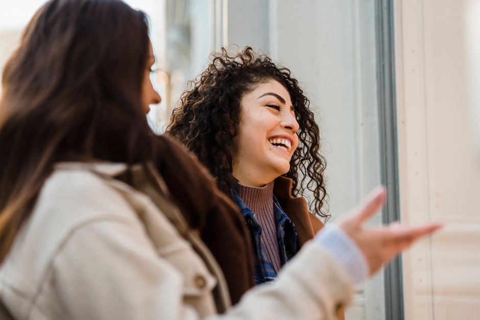 Side view of happy young multiethnic female best friends with long dark hairs in casual clothes laughing while looking at showcase of fashion store on city street